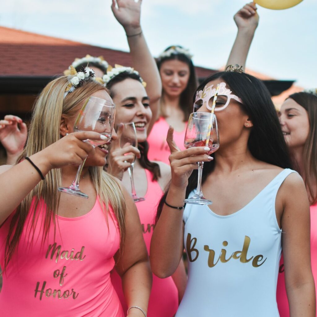 Un grupo de mujeres celebrando, vestidas con trajes de baño a juego. Una lleva un traje blanco con la palabra "Bride" y otra lleva uno rosa con la palabra "Maid of Honor". Todas están sonriendo y levantando copas de vino.