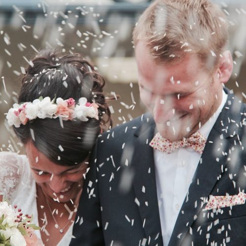 Novia y novio sonriendo mientras les lanzan arroz a la salida de la ceremonia. La novia lleva una corona de flores y el novio un traje oscuro con pajarita.