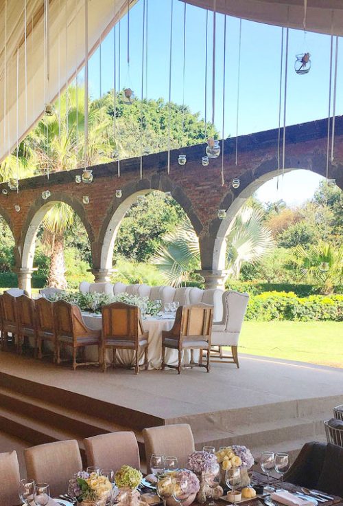 Mesa de banquete en un jardín con una estructura de arcos de piedra y techado. La mesa está decorada con manteles blancos y flores, y hay sillas elegantes alrededor.